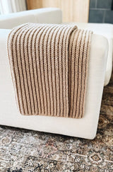 Ribbed knit throw in a tan yarn draped over the arm of an off-white couch, in a living room.  thumbnail