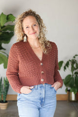Front view of woman wearing warm rusty-red crochet cardigan buttoned closed, cardigan ends at waist, with blue jeans.  thumbnail