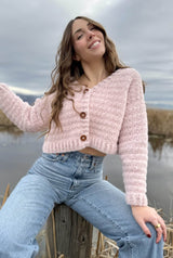 Woman sitting on wooden stump in blue jeans, with fuzzy pink cropped cardigan with three buttons.  thumbnail