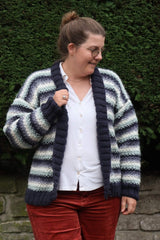 Woman wearing a striped knit cardigan in white, navy, mauve, and seafoam, over white shirt and cranberry pants, stands in front of evergreen hedge.  thumbnail