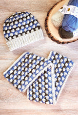 Crochet hat and matching cowl lay flat on wooden table, with yarn skeins stacked on log slice. Hat and cowl are crocheted in cream, denim blue, and dark olive green.  thumbnail