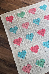 White crochet blanket with multi color heart blocks laid on wooden floor.  thumbnail