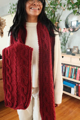 Smiling woman holds end of cranberry red crochet scarf in hand, with other end hanging free.  thumbnail