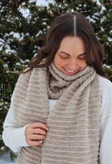 Woman wears a beige knit scarf with textured ridges. She wears a white sweater and stands in front of an evergreen bush dusted with snow.  thumbnail