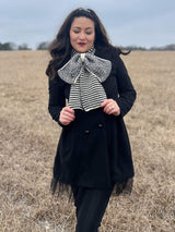 Woman wears cream and white bow scarf over black coat and tulle skirt, standing in winter field.  thumbnail