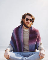 Bearded man in beige sweater and pale blue pants sits cross-legged, wearing knit wrap in shades of purple.  thumbnail