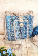 Blue and tan crochet bag sitting upright on wooden bench. Two balls of yarn and a bouquet of flowers are in the foreground.  thumbnail