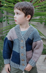 Young boy stands in front of greenery. He wears a patchwork knit cardigan in shades of blue, buttoned up the front.  thumbnail