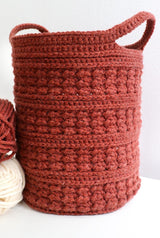 Red-orange crochet basket with bands of different textures and two handles sits against white backdrop.  thumbnail