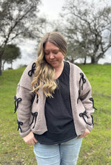 Woman wears a tan cardigan with 3-D black bows over a black shirt and jeans. She stands in a green grassy field.  thumbnail