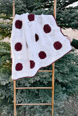 Crochet blanket made of white squares with a dark red circle in the center of each. Blanket hangs on wooden ladder balanced against evergreen tree.  thumbnail