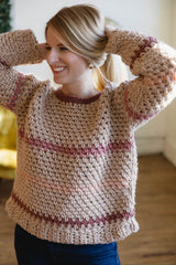 Front view of woman with hands up in hair, wearing tan sweater with multi-pink stripes.   thumbnail