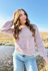 Woman with long brunette hair stands in front of frozen pond, wearing blush pink crochet sweater thumbnail