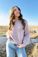 Woman wearing blush pink textured crochet sweater, standing in brown field with frozen pond in background thumbnail