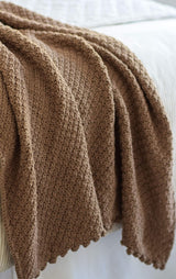Warm brown crochet blanket in textured stitch pattern draped over white bedding thumbnail