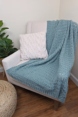 Sky blue crochet blanket draped over cream armchair, with a commercially knit pillow and a fake plant nearby.  thumbnail