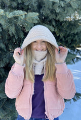 Woman wears combination hood/cowl in cream color. She has a pink coat and a purple sweater on, and stands in front of a pine tree in the snow. thumbnail