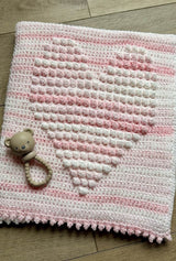 Folded baby blanket showing heart motif made with popcorn stitches. Yarn is in shades of soft pink.  thumbnail