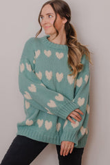 Woman stands in front of white backdrop wearing a seafoam green sweater with cream hearts all over it.  thumbnail