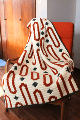 Crochet afghan draped over rust colored chair. Afghan is made with geometric motifs in rust and dark green on a cream background.  thumbnail