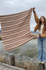 Woman in tan jacket, white shirt and blue jeans stands on a wooden post, holding afghan to her side over a frozen wetland. Afghan is in beige with textured stripes of purple, red, and brown.  thumbnail