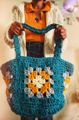 Woman hods large crochet bag toward camera. Bag body is made of large granny squares in cream, mustard, grey, and teal. Straps are long enough to go over shoulder.  thumbnail
