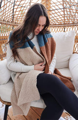 Woman sits in wicker chair wearing white top, black leggings, and a large color block scarf. Scarf is knit in a textured stitch and has sections of beige, teal, and warm brown.  thumbnail