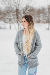 Woman standing in snowy yard with trees in the background, wearing white tee and blue jeans. Over that she wears an open grey crochet cardigan, with her hands in the pockets.  thumbnail