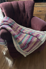 Crocheted afghan in shades of purple aqua, and tan. It lays on a dark eggplants armchair, sitting on a wooden floor.  thumbnail