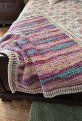 Crochet afghan in self-striping yarn in shades of purple, pink, pale yellow, and turquoise. It's laid on a bed with dark wooden frame and a floral bedspread.  thumbnail