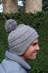 Man wears knit hat in pale grey. The hat has a pompom, a sideways cabled brim, and shapes earflaps. He wears a steel blue puffer coat and stands in front of an evergreen hedge.  thumbnail