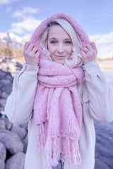 Woman wears a fluffy pink knit scarf with fringe. The scarf is wrapped around and worn as a hood. She wears it over a cream coat and is standing on a rocky shoreline.  thumbnail