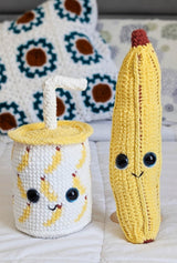 Two crocheted stuffed toys sitting on a white bedspread, with decorative pillows in the background. One toy is a banana, with large eyes and a brown stem at top. The other toy is a smoothie cup with a banana pattern. It has a yellow lid and a white straw. thumbnail