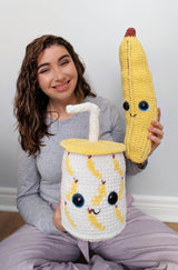 Woman seated cross-legged holds smoothie cup and stuffed banana. The cup is about 12 inches tall, and the banana is 16 inches tall.  thumbnail