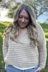 Woman wears a beige crochet mesh pullover over a white t-shirt and jeans. She stands in front of a grassy hill with a tree on top. thumbnail