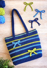 Crochet tote bag laid flat on pale wood background. Bag is in navy blue with stripes of paler blue and green-yellow. It has two bows and handles at the top.  thumbnail