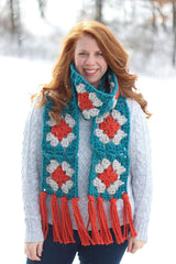 Woman in grey sweater and dark jeans stands in the snow. She wears a granny square scarf in dark teal, pale grey, and red-orange, with fringe on each end.  thumbnail