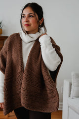 Woman wears a brown ruana style poncho over a cream turtleneck, with black pants. She stands in a living room with a white couch and a wooden mantel visible behind.  thumbnail