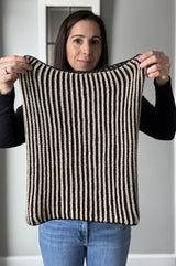 Woman holds unfolded cowl toward camera, showing columns of tan stitches on a black background.  thumbnail