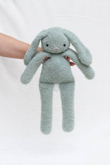 Sage green bunny toy help under the arms with legs hanging down. It has long floppy ears, black eyes, and a pink nose.  thumbnail