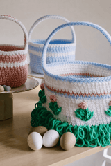 Three crochet Easter baskets sit on light wooden table. The large basket is in white with blue and pink stripes. It has flowers on the side and green loop texture at the bottom to mimic grass.  thumbnail