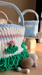 Close up of larger basket showing grass trim and floral appliques. Some fake eggs and candy eggs are nearby. In the background is a smaller blue basket.  thumbnail
