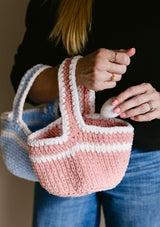 Woman in black top and blue jeans hold two smaller crochet baskets on her arm, putting an egg into the pink & white one.  thumbnail