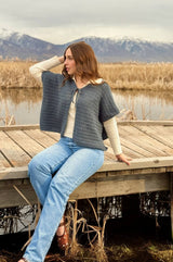 Woman sits on a wooden dock in a wetland, with snow-capped mountains in the distance. She wears jeans, a cream long sleeve top, and a grey crocheted cape cardigan.  thumbnail