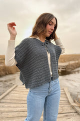Woman stands on a wooden boardwalk in blue jeans. She ahs her arms raised and is wearing a crochet cropped cape cardigan in grey.  thumbnail