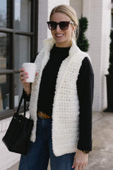 Woman stands on a sidewalk in a shopping district. She wears jeans, a black long sleeve shirt, sunglasses, and has a small purse over the arm carrying a coffee cup. Over the top she wears a white crochet fluffy open vest with a stand up collar.  thumbnail