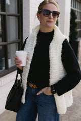 Woman stands on sidewalk with hand in jeans pocket, holding coffee up in other hand. She wears a white crochet fluffy vest over a black long sleeve top and jeans. A purse is hanging in the bend of her right arm.  thumbnail