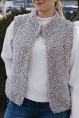 Cropped view of woman in a white long sleeve top and jeans, standing in front of a hedge. She wears a pale brown knit textured vest. The vest is open and has a standing collar.  thumbnail