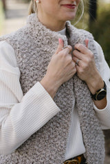 Close up of a woman in a white top pulling closed an open front vest  The vest is knit in a fluffy tan yarn.  thumbnail