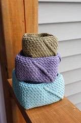 Three crochet baskets sit on a wooden porch railing. The baskets are arranged from largest to smallest and are shown in aqua, lilac, and olive green.  thumbnail